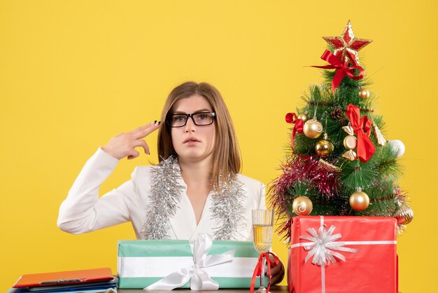 Vista frontal doctora sentada delante de la mesa con regalos y árbol sobre fondo amarillo con árbol de navidad y cajas de regalo