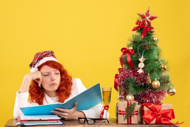 Vista frontal doctora sentada detrás de la mesa leyendo documentos en el escritorio amarillo con árbol de navidad y cajas de regalo