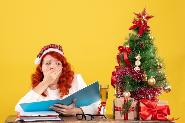 Vista frontal doctora sentada detrás de la mesa leyendo documentos sobre fondo amarillo con árbol de navidad y cajas de regalo