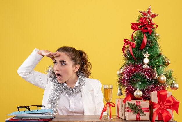 Vista frontal doctora sentada detrás de la mesa mirando a distancia sobre fondo amarillo con árbol de navidad y cajas de regalo