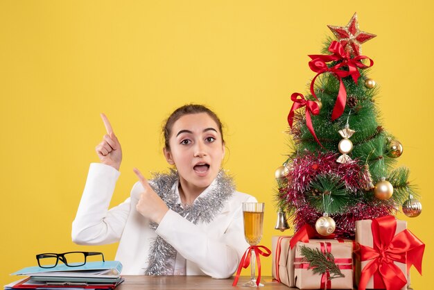 Vista frontal doctora sentada detrás de la mesa con regalos de Navidad y árbol sobre fondo amarillo