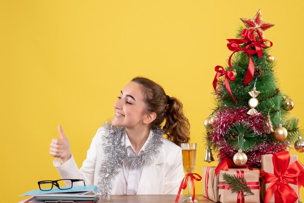 Vista frontal doctora sentada detrás de la mesa con regalos de Navidad y árbol sobre fondo amarillo