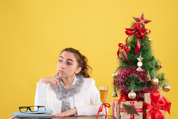 Vista frontal doctora sentada detrás de la mesa con regalos de Navidad y árbol sobre fondo amarillo