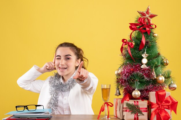 Vista frontal doctora sentada detrás de la mesa con regalos de Navidad y árbol sobre fondo amarillo