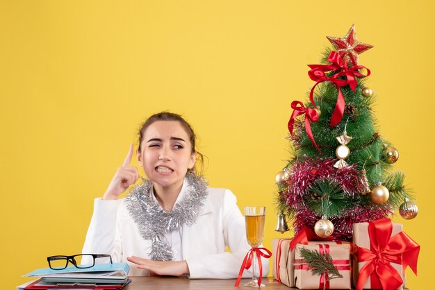 Vista frontal doctora sentada detrás de la mesa con regalos de Navidad y árbol sobre fondo amarillo