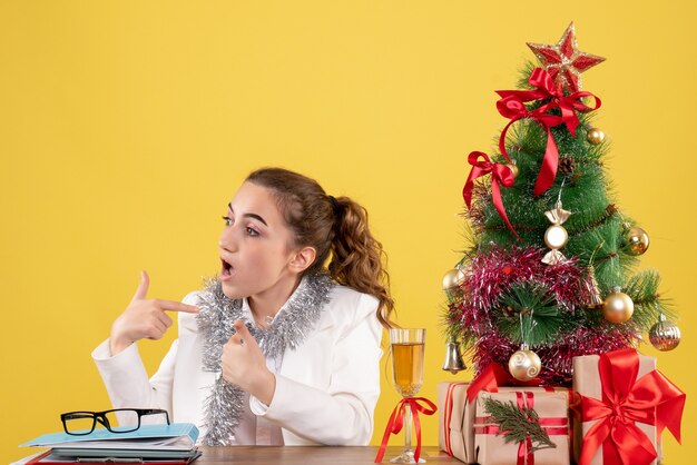 Vista frontal doctora sentada detrás de la mesa con regalos de Navidad y árbol sobre fondo amarillo