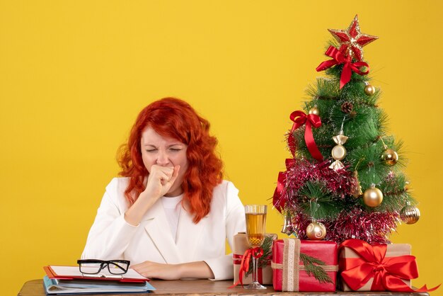 Vista frontal doctora sentada detrás de la mesa con regalos de navidad bostezando sobre fondo amarillo