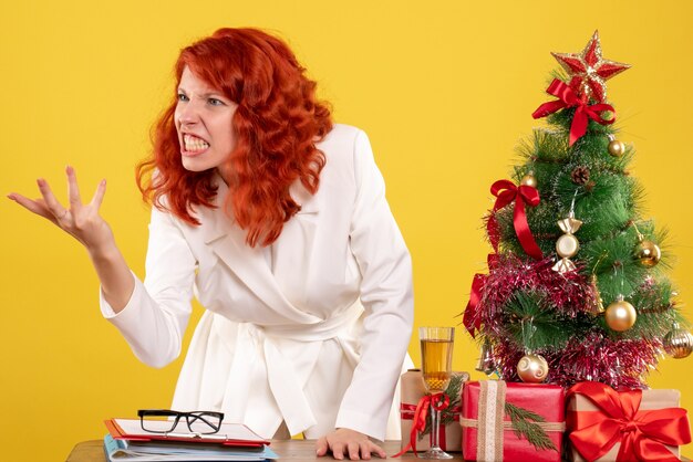 Vista frontal doctora sentada detrás de la mesa con regalos de navidad discutiendo sobre fondo amarillo con árbol de navidad y cajas de regalo