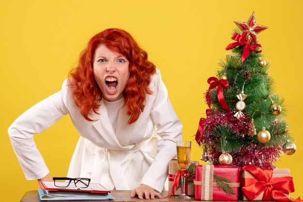 Vista frontal doctora sentada detrás de la mesa con regalos de navidad enojado sobre fondo amarillo con árbol de navidad y cajas de regalo