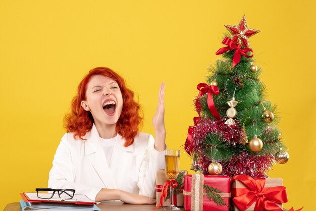 Vista frontal doctora sentada detrás de la mesa con regalos de navidad levantando su mano sobre fondo amarillo