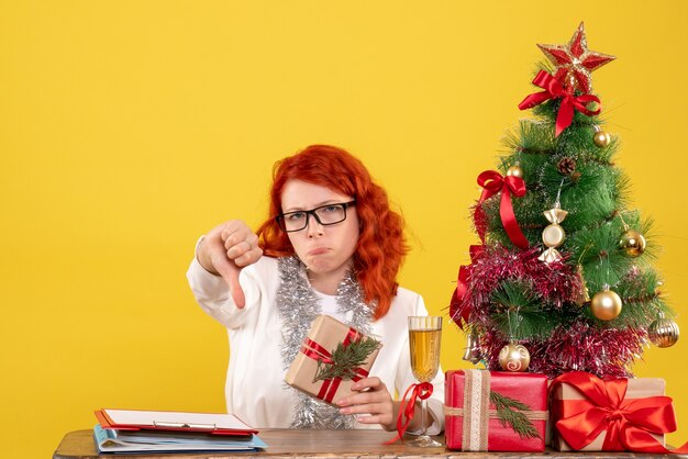 Vista frontal doctora sentada detrás de la mesa con regalos de navidad sintiéndose triste sobre fondo amarillo