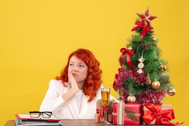 Vista frontal doctora sentada detrás de la mesa con regalos de navidad sobre fondo amarillo