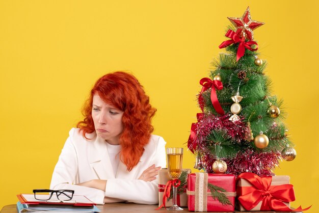 Vista frontal doctora sentada detrás de la mesa con regalos de navidad sobre fondo amarillo