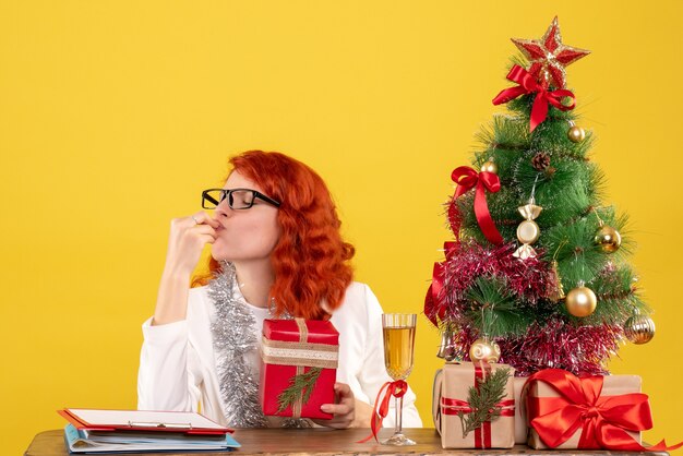 Vista frontal doctora sentada detrás de la mesa con regalos de navidad sobre fondo amarillo