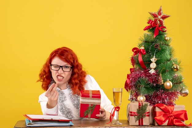 Vista frontal doctora sentada detrás de la mesa con regalos de navidad sobre fondo amarillo