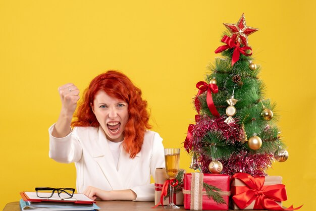 Vista frontal doctora sentada detrás de la mesa con regalos de navidad sobre fondo amarillo