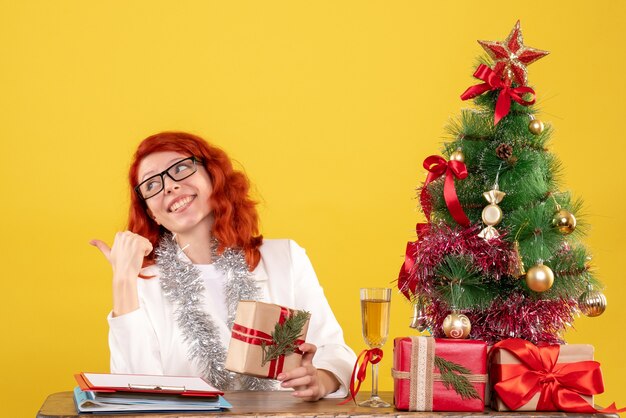 Vista frontal doctora sentada detrás de la mesa con regalos de navidad sobre fondo amarillo