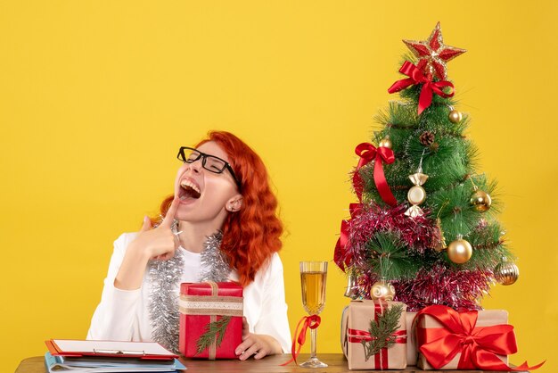 Vista frontal doctora sentada detrás de la mesa con regalos de navidad sobre fondo amarillo