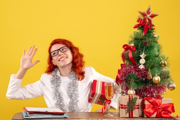 Vista frontal doctora sentada detrás de la mesa con regalos de navidad sobre fondo amarillo