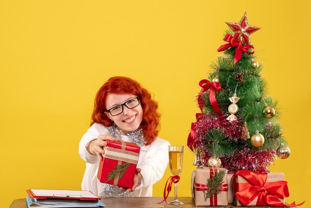 Vista frontal doctora sentada detrás de la mesa con regalos de navidad sobre fondo amarillo