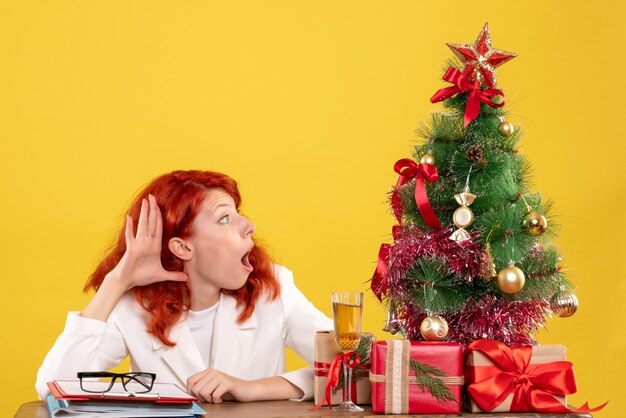 Vista frontal doctora sentada detrás de la mesa con regalos de navidad sobre fondo amarillo