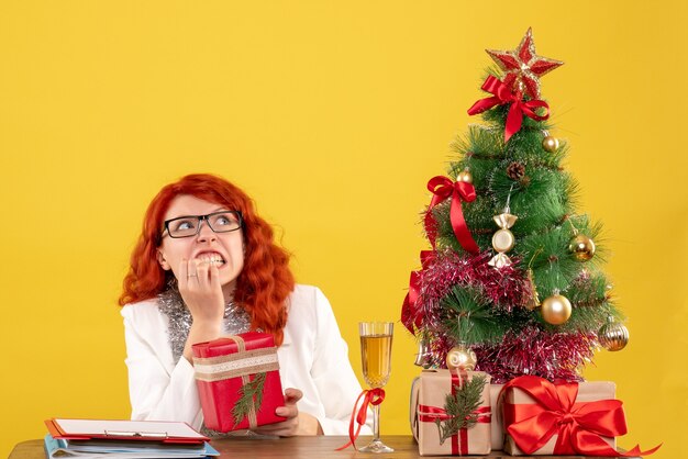 Vista frontal doctora sentada detrás de la mesa con regalos de navidad sobre fondo amarillo