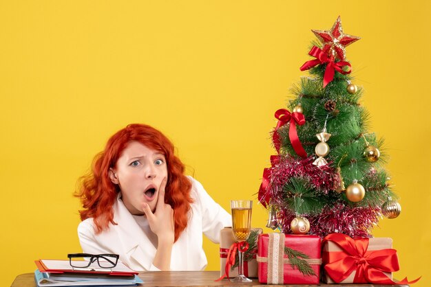 Vista frontal doctora sentada detrás de la mesa con regalos de navidad sobre fondo amarillo