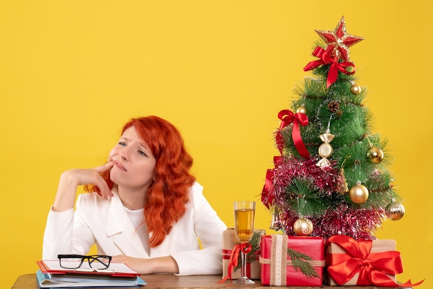 Vista frontal doctora sentada detrás de la mesa con regalos de navidad sobre fondo amarillo