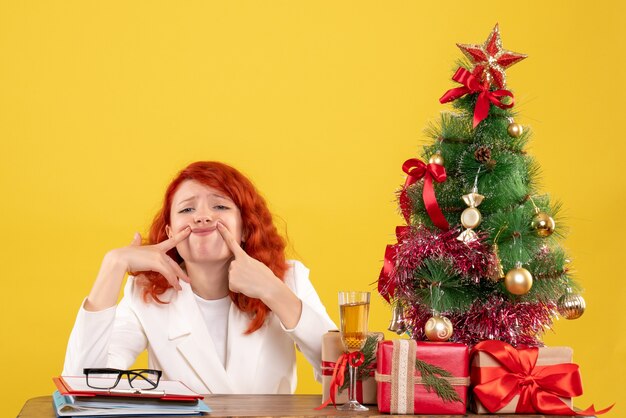 Vista frontal doctora sentada detrás de la mesa con regalos de navidad sobre fondo amarillo