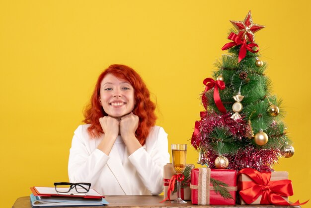 Vista frontal doctora sentada detrás de la mesa con regalos de navidad sonriendo sobre fondo amarillo