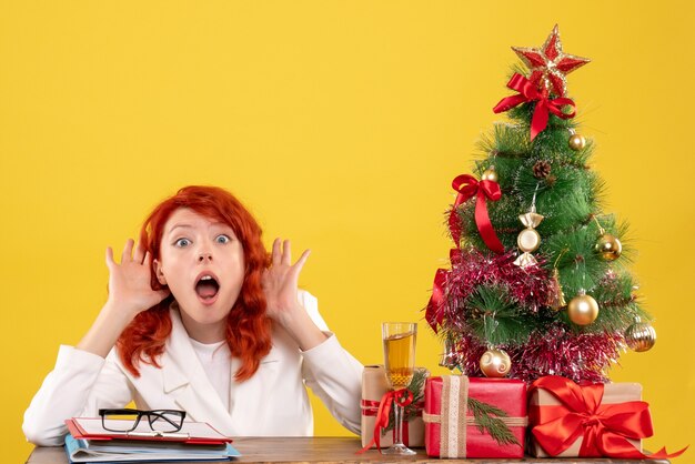 Vista frontal doctora sentada detrás de la mesa con regalos de navidad sorprendidos sobre fondo amarillo