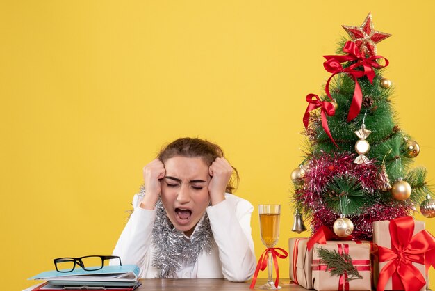 Vista frontal doctora sentada detrás de la mesa con regalos de Navidad y sufrimiento de árboles sobre fondo amarillo