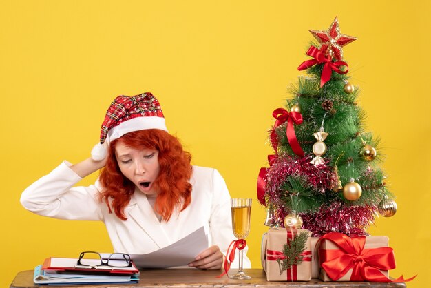 Vista frontal doctora sentada detrás de la mesa con regalos sobre un fondo amarillo con árbol de navidad y cajas de regalo