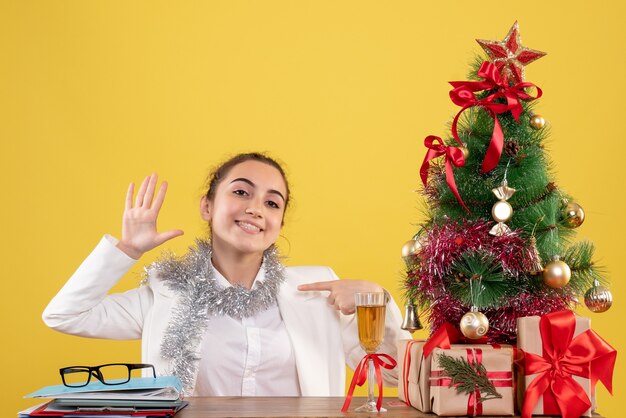 Vista frontal doctora sentada detrás de la mesa sobre fondo amarillo con árbol de navidad y cajas de regalo