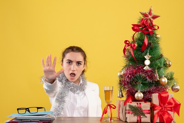 Vista frontal doctora sentada detrás de la mesa sobre fondo amarillo con árbol de navidad y cajas de regalo