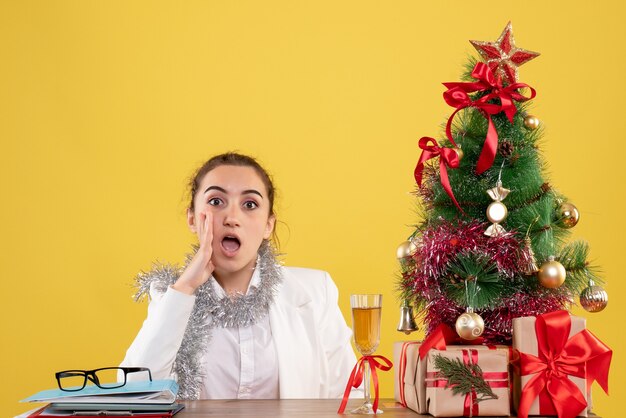 Vista frontal doctora sentada detrás de la mesa sobre fondo amarillo con árbol de navidad y cajas de regalo