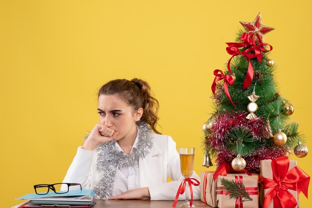 Vista frontal doctora sentada detrás de su mesa con cara estresada sobre fondo amarillo con árbol de navidad y cajas de regalo