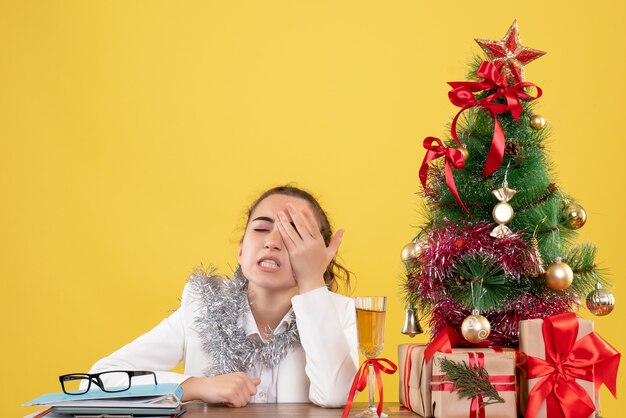 Vista frontal doctora sentada detrás de su mesa destacó sobre fondo amarillo con árbol de navidad y cajas de regalo