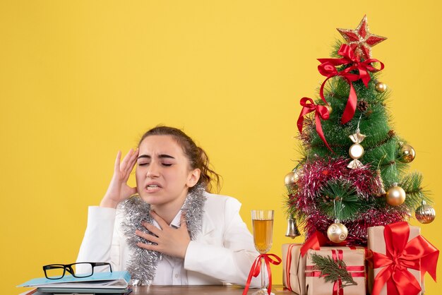 Vista frontal doctora sentada detrás de su mesa con dolor de cabeza sobre fondo amarillo con árbol de navidad y cajas de regalo
