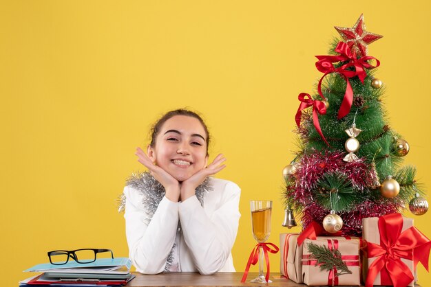 Vista frontal doctora sentada detrás de su mesa emocionada sobre fondo amarillo con árbol de navidad y cajas de regalo