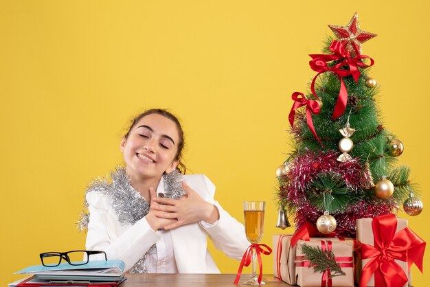 Vista frontal doctora sentada detrás de su mesa encantada sobre fondo amarillo con árbol de navidad y cajas de regalo