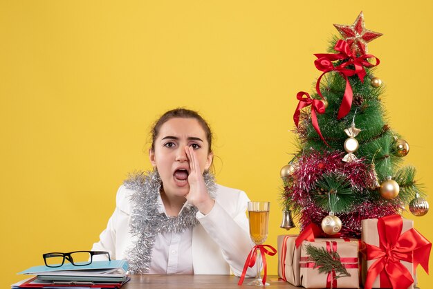 Vista frontal doctora sentada detrás de su mesa llamando sobre fondo amarillo con árbol de navidad y cajas de regalo