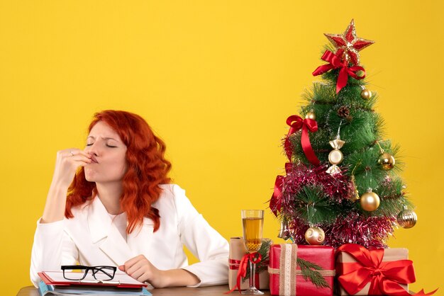 Vista frontal doctora sentada detrás de su mesa con regalos de navidad y árbol en escritorio amarillo