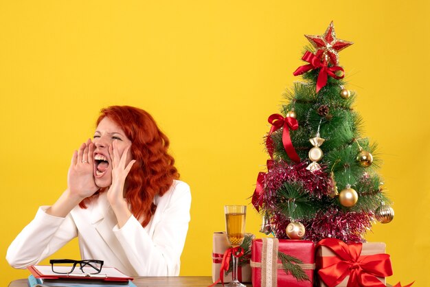 Vista frontal doctora sentada detrás de su mesa con regalos de Navidad y árbol llamando sobre fondo amarillo