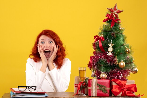 Vista frontal doctora sentada detrás de su mesa con regalos de navidad y árbol sobre fondo amarillo