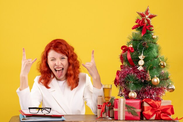 Vista frontal doctora sentada detrás de su mesa con regalos de navidad y árbol sobre fondo amarillo