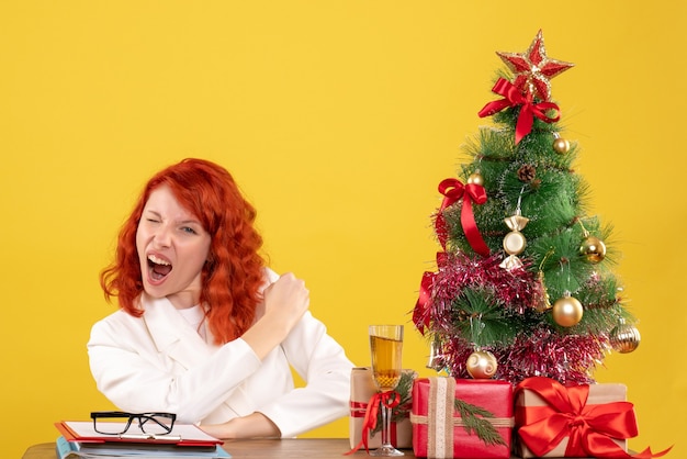 Vista frontal doctora sentada detrás de su mesa con regalos de navidad y árbol sobre fondo amarillo