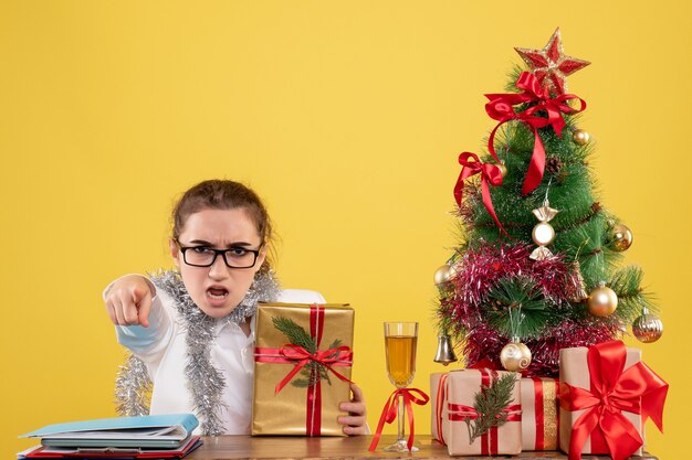 Vista frontal doctora sentada detrás de su mesa con regalos de Navidad y árbol sobre fondo amarillo