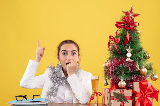 Vista frontal doctora sentada detrás de su mesa sobre fondo amarillo con árbol de navidad y cajas de regalo