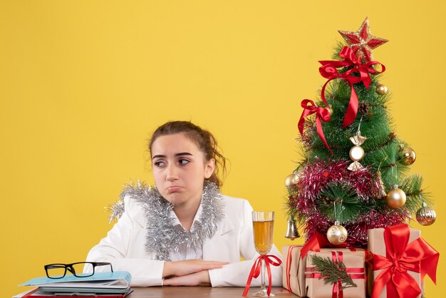 Vista frontal doctora sentada detrás de su mesa sobre fondo amarillo con árbol de navidad y cajas de regalo